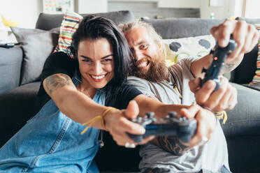 Bärtiger, tätowierter Mann mit langen brünetten Haaren und Frau mit langen braunen Haaren sitzen auf einem Sofa und spielen lächelnd ein Konsolenspiel. - CUF56063