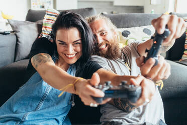 Bearded tattooed man with long brunette hair and woman with long brown hair sitting on a sofa, smiling while playing console game. - CUF56063