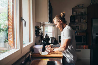 Bearded tattooed man with long brunette hair standing in a kitchen, preparing food. - CUF56066