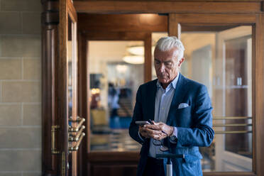 Ein älterer Geschäftsmann steht in der Hotellobby und schaut auf sein Mobiltelefon. - CUF56080