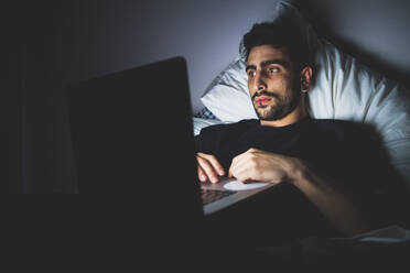 Bärtiger junger Mann liegt nachts im Bett und schaut auf seinen Laptop. - CUF56093