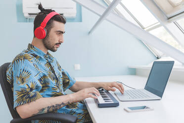 Young bearded man sitting at a table, wearing headphones, typing on laptop and playing keyboard. - CUF56101