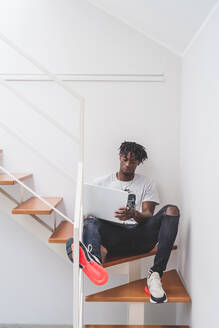 Young man with short dreadlocks sitting on staircase, typing on laptop. - CUF56105