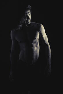 Gutaussehender Mann ohne Hemd mit langen Haaren steht in einem dunklen Raum und schaut weg - ADSF01261