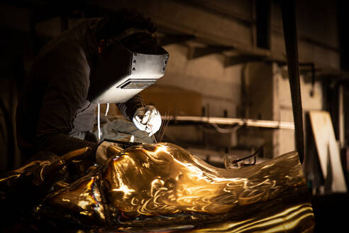 Side view of male in welding helmet working with metal product in obscurity on little foundry - ADSF01274