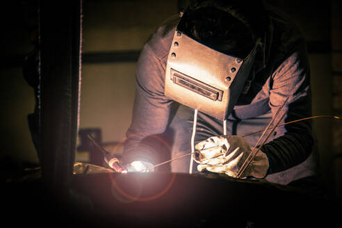 Side view of male in welding helmet working with metal product in obscurity on little foundry - ADSF01275