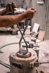 Die Hände eines Mannes mit einer Hebezange, der ein Metallprodukt aus einem Schmelztiegel in einer kleinen Gießerei nimmt - ADSF01277
