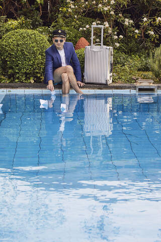 Young man floating paper boats on swimming pool while sitting with suitcase against plants stock photo