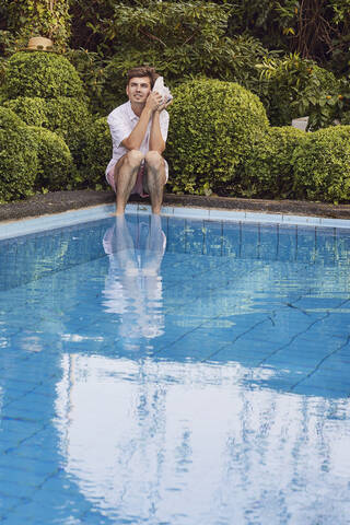 Young man listening through seashell while sitting at poolside against plants stock photo