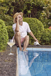 Nachdenklicher junger Mann mit Hut sitzt mit Muschelschale am Pool - UKOF00008
