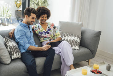 Ein glückliches Paar benutzt ein digitales Tablet, während es auf dem Sofa im Wohnzimmer eines Penthouses sitzt - EHF00541