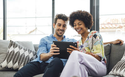 Glückliches multiethnisches Paar, das ein digitales Tablet benutzt, während es auf dem Sofa im Penthouse sitzt - EHF00550