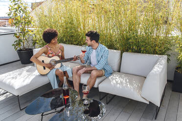 Happy woman playing guitar for man drinking red wine on sofa at penthouse patio - EHF00559