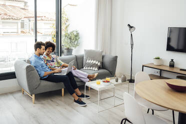 Multi-ethnic couple using laptop while sitting on sofa in modern penthouse - EHF00560