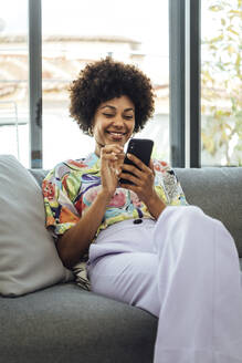 Glückliche Frau im mittleren Erwachsenenalter, die ein Smartphone benutzt, während sie auf dem Sofa in einem modernen Penthouse sitzt - EHF00564