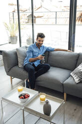 Mittlerer erwachsener Mann, der ein Smartphone benutzt, während er auf dem Sofa im Wohnzimmer eines modernen Penthouses sitzt - EHF00567