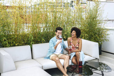 Cheerful couple having red wine while sitting on sofa at penthouse patio - EHF00600