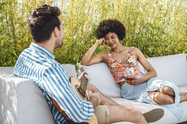 Woman holding red wine glass while looking at man playing guitar on sofa at penthouse patio - EHF00603