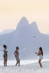 Ein junger schwarzer Brasilianer spielt Fußball mit zwei Freundinnen am Strand von Ipanema mit den Dois Irmaos Bergen in der Ferne, Rio de Janeiro, Brasilien, Südamerika - RHPLF16164