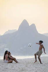 Ein junger schwarzer Brasilianer spielt Fußball mit zwei Freundinnen am Strand von Ipanema mit den Dois Irmaos Bergen in der Ferne, Rio de Janeiro, Brasilien, Südamerika - RHPLF16165