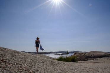Sun shining over teenage girl walking alone along rocky shore - LBF03164