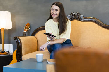 Happy young woman holding smart phone while sitting on sofa in modern cafe - GIOF08502