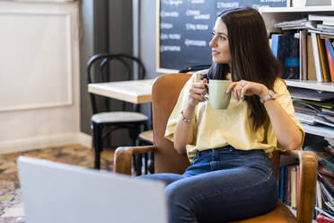 Thoughtful young woman holding coffee mug while sitting on armchair in coffee shop - GIOF08517