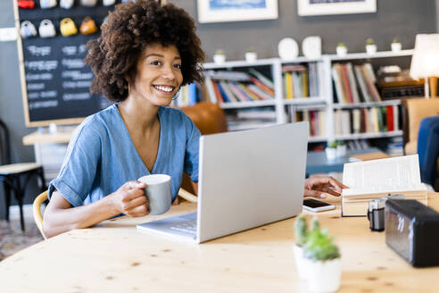 Nachdenkliche Frau, die eine Kaffeetasse hält, während sie mit einem Laptop an einem Kaffeetisch sitzt - GIOF08519