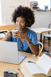 Thoughtful woman holding coffee mug while sitting with laptop at coffee shop - GIOF08520