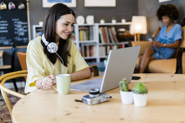 Glückliche Frau mit Laptop in einem Café mit einem Freund im Hintergrund - GIOF08522