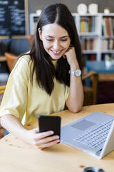 Happy woman using smart phone while sitting with laptop at coffee shop - GIOF08523