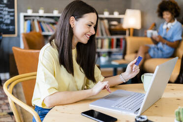 Happy woman using credit card and laptop while shopping online in cafe - GIOF08525