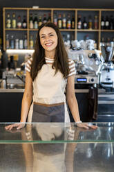 Happy female owner standing at counter in coffee shop - GIOF08551