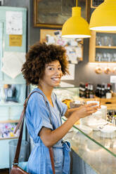 Glückliche junge Frau, die eine Kaffeetasse hält, während sie am Tresen eines Cafés steht - GIOF08556