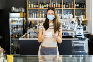 Beautiful owner wearing mask while holding coffee cup and saucer in cafe - GIOF08585