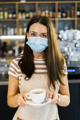 Beautiful young owner wearing mask while holding coffee cup and saucer in cafe - GIOF08586