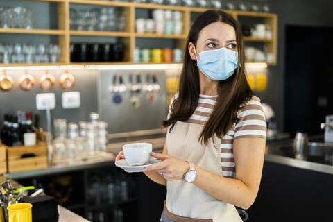 Nachdenklicher junger Besitzer trägt eine Maske und hält eine Kaffeetasse mit Untertasse in einem Café, lizenzfreies Stockfoto