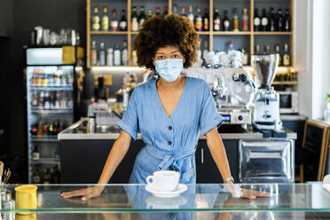 Young female owner wearing mask standing at cafe counter - GIOF08588