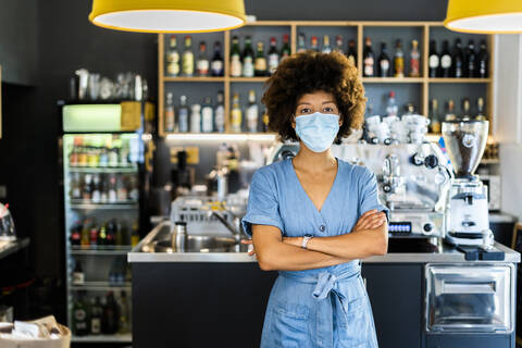 Junge Frau mit Maske steht mit verschränkten Armen in einem Café, lizenzfreies Stockfoto
