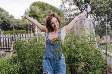 Cheerful woman with arms raised standing against plants in vegetable garden - EBBF00426