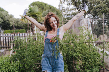 Fröhliche Frau mit erhobenen Armen, die gegen Pflanzen im Gemüsegarten steht - EBBF00426
