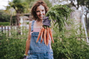 Smiling mid adult woman holding carrots while standing in vegetable garden - EBBF00427