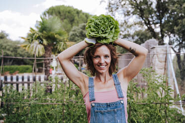 Smiling woman holding lettuce on head while standing in vegetable garden - EBBF00429