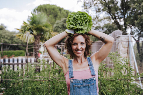 Lächelnde Frau hält Salat auf dem Kopf, während sie im Gemüsegarten steht - EBBF00429