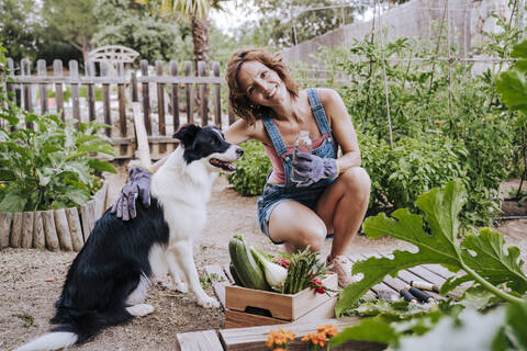 Smiling woman holding water bottle crouching by border collie in vegetable garden stock photo