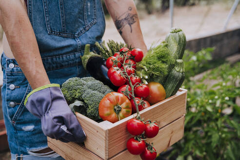 Close-up of woman holding wooden crate with various vegetables while standing in community garden - EBBF00435