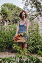 Smiling woman holding various vegetables in crate while standing against plants at community garden - EBBF00436