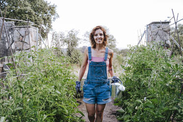 Lächelnde Frau, die eine Gießkanne hält, während sie inmitten von Pflanzen im Gemüsegarten steht - EBBF00449