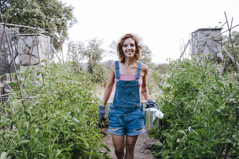 Smiling woman holding watering can while standing amidst plants in vegetable garden - EBBF00449