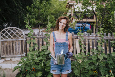 Smiling woman holding watering can while standing in vegetable garden - EBBF00451